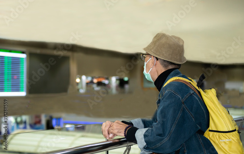 Man with yellow backpack, brown hat and medical face mask wearing due to covid-19 or coronavirus outbreak or pandemic standing with arms on the iron rail for look around in the airport on holiday.
