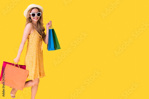 Picture of a shocked young brunette woman in yellow summer dress wearing sunglasses posing with shopping bags and orange background.