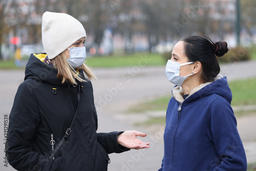 Two women in medical protective masks communicate in street. Safe walks in coronavirus pandemic concept