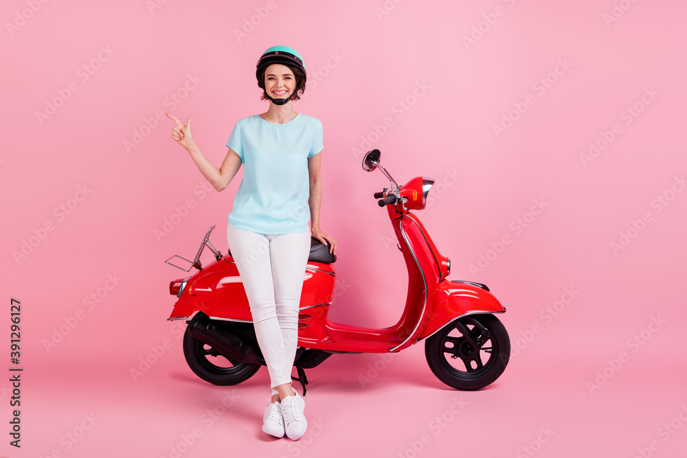 Photo of positive pretty young lady stand moped point empty space wear white trousers footwear blue slam t-shirt isolated on pink background Photo of positive pretty young lady stand moped point empty space wear white trousers footwear blue slam t-shirt isolated on pink background