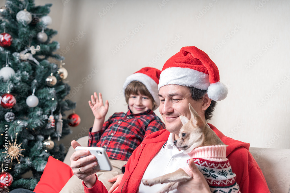 Father with child and puppy dog in Santa hats having a video call on Christmas day at smartphone, sitting on a couch in the living room with Christmas tree at home. Father with child and puppy dog in Santa hats having a video call on Christmas day at smartphone, sitting on a couch in the living room with Christmas tree at home.