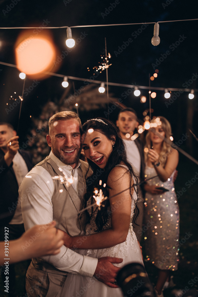 The bride and groom congratulate each other against the background of a flower arch. Evening wedding. Fireworks and lights.