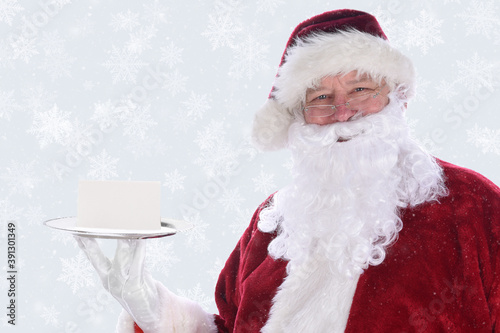 Santa Claus holding a silver platter with a blank note card, over a snow flake background with snow effect. © Steve Cukrov