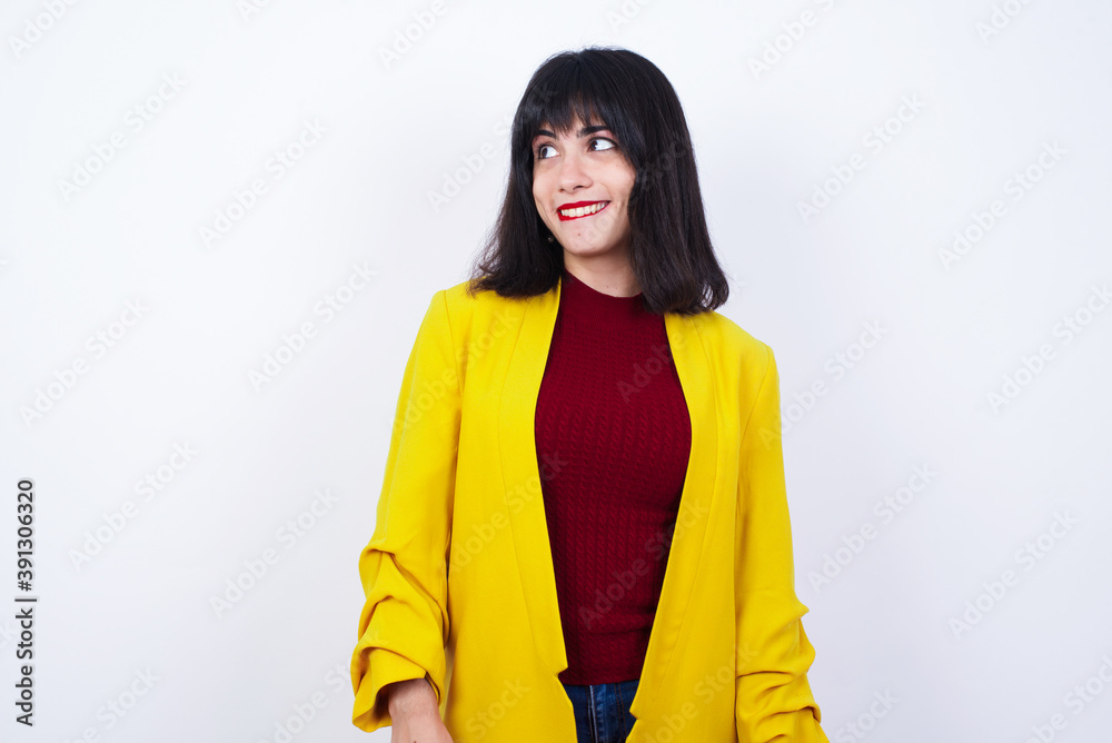 Caucasian woman standing against white background wearing yellow blazer with thoughtful expression, looks away keeps hands down bitting his lip thinks about something pleasant. Caucasian woman standing against white background wearing yellow blazer with thoughtful expression, looks away keeps hands down bitting his lip thinks about something pleasant.