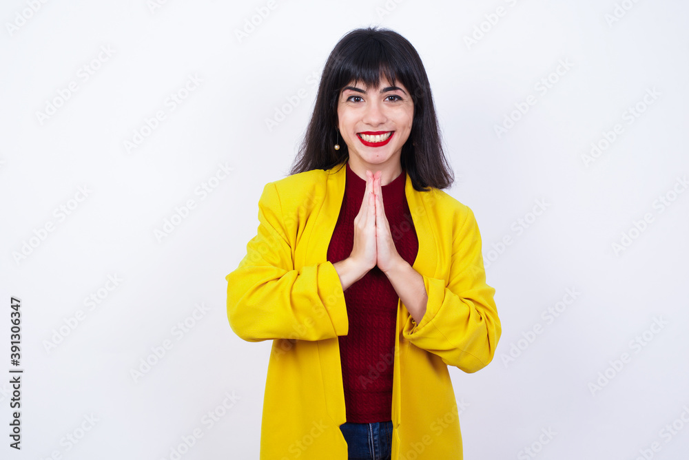 Caucasian woman standing against white background wearing yellow blazer keeps palms together, has pleased expression. Glad attractive male makes request, pleads for mercy. Hopeful young adult. Caucasian woman standing against white background wearing yellow blazer keeps palms together, has pleased expression. Glad attractive male makes request, pleads for mercy. Hopeful young adult.