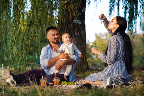 Young parents with baby having picnic under willow trees near river. Mother blowing bubbles and father holding little child. Concept of fun