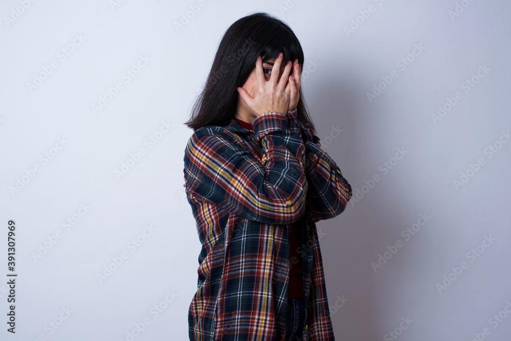 Beautiful young Caucasian woman standing against white background covering face with hands and peering out with one eye between fingers. Scared from something or someone. Beautiful young Caucasian woman standing against white background covering face with hands and peering out with one eye between fingers. Scared from something or someone.