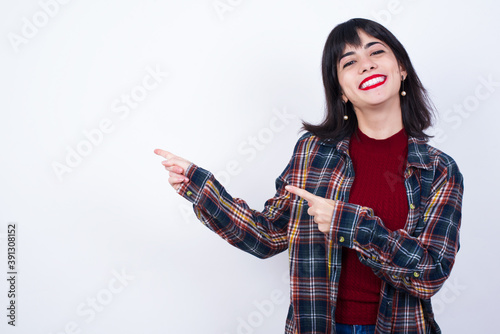 Beautiful young Caucasian woman standing against white background with positive expression, indicates with fore finger at blank copy space for your promotional text or advertisement.