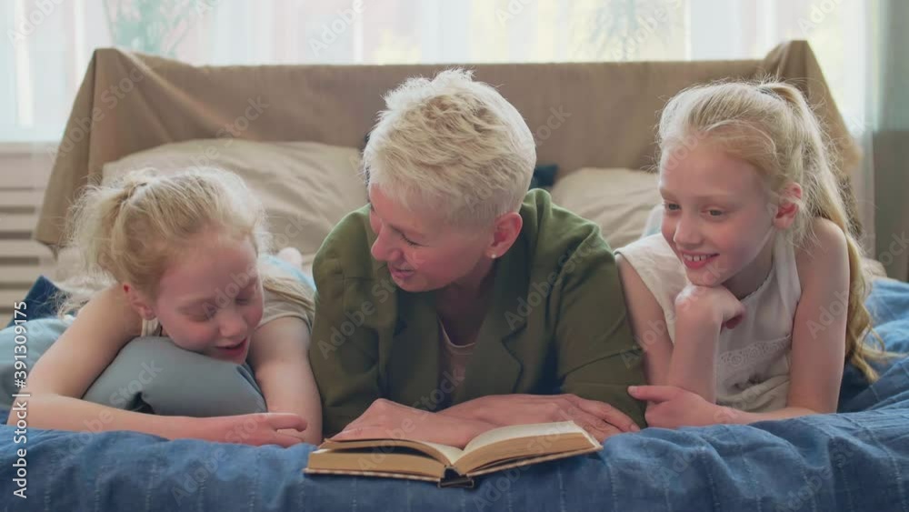 Medium close-up POV of good-looking Caucasian senior woman lying on bed reading book to cheerful blond-haired twin granddaughters. Family looking up and smiling on cameraMedium close-up POV of good-lo Medium close-up POV of good-looking Caucasian senior woman lying on bed reading book to cheerful blond-haired twin granddaughters. Family looking up and smiling on cameraMedium close-up POV of good-lo