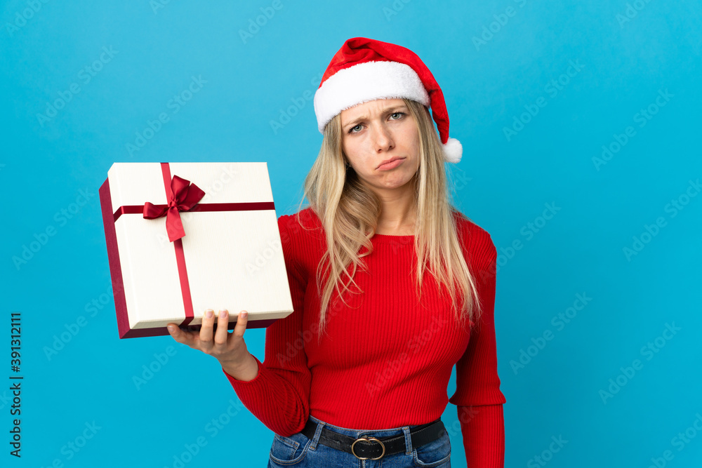 Woman with christmas hat holding a present isolated on blue background with sad expression Woman with christmas hat holding a present isolated on blue background with sad expression