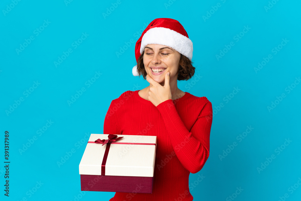 English girl with christmas hat holding a present isolated on blue background looking to the side and smiling English girl with christmas hat holding a present isolated on blue background looking to the side and smiling