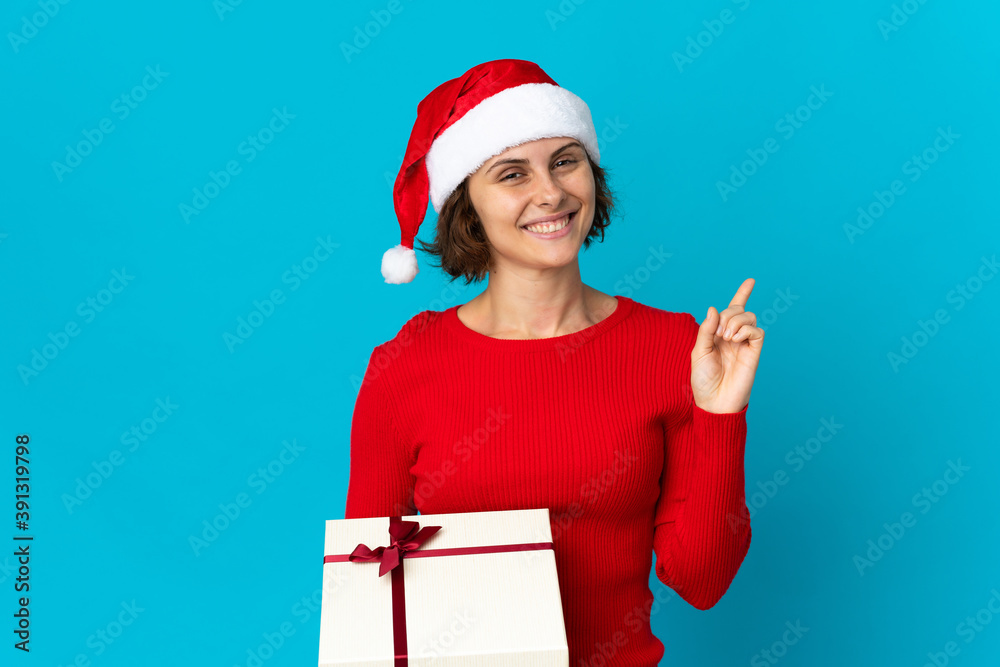 English girl with christmas hat holding a present isolated on blue background showing and lifting a finger in sign of the best English girl with christmas hat holding a present isolated on blue background showing and lifting a finger in sign of the best