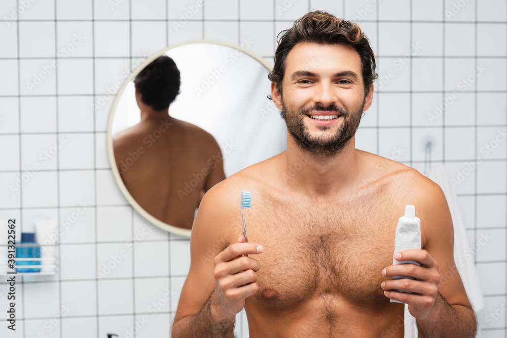 Cheerful shirtless man looking at camera while holding toothpaste and toothbrush in bathroom Cheerful shirtless man looking at camera while holding toothpaste and toothbrush in bathroom