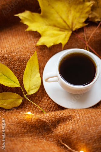 A cup of coffee on an orange sweater, yellow leaves, lights in the background. Warm autumn.