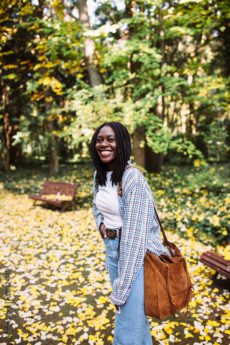 Portrait of a pretty black woman in stylish casual shirt with brown bag walking in an autumn park with colorful yellow fall leaves