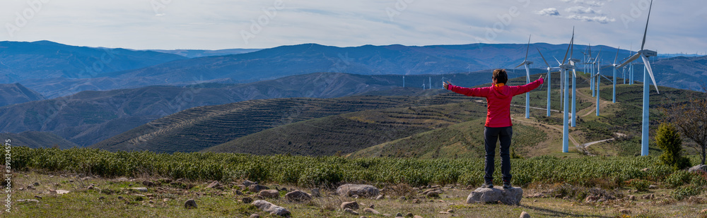 woman in red mountain clothes, with open arms in the form of a cross, enjoys her freedom and happiness and contemplates a wind farm and the mountains