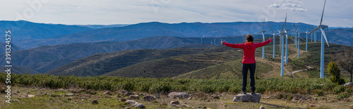 woman in red mountain clothes, with open arms in the form of a cross, enjoys her freedom and happiness and contemplates a wind farm and the mountains