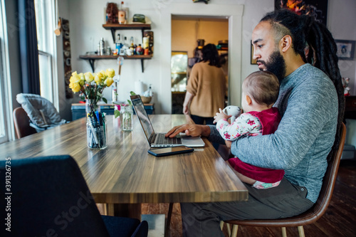 Dad feeding baby while working from home on laptop computer at dining room table
