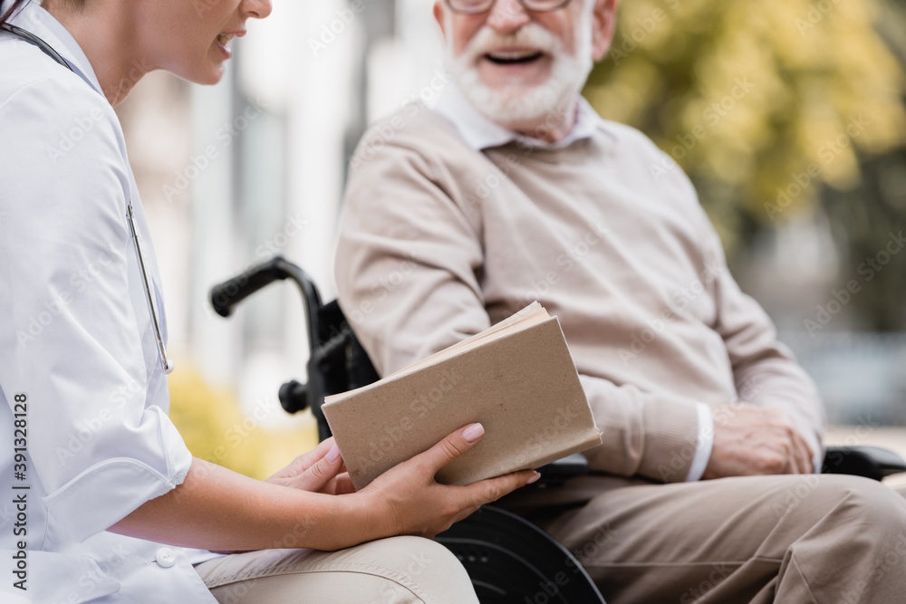 Fototapeta premium cropped view of geriatric nurse reading book to aged man on blurred background