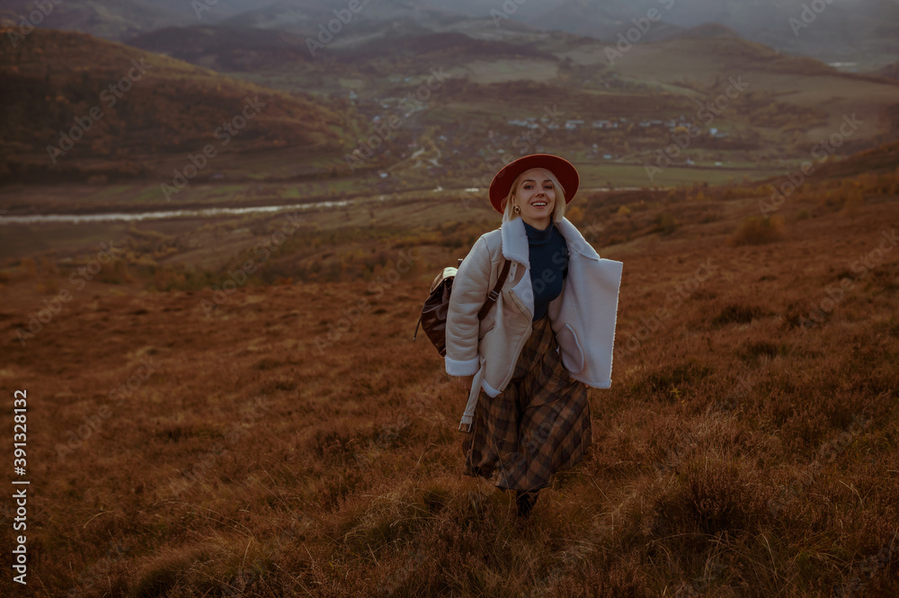 Outdoor autumn fashion portrait of young elegant happy smiling woman wearing trendy hat, white faux sheepskin jacket, checkered skirt, walking in nature, mountain landscape. Copy, empty space for text