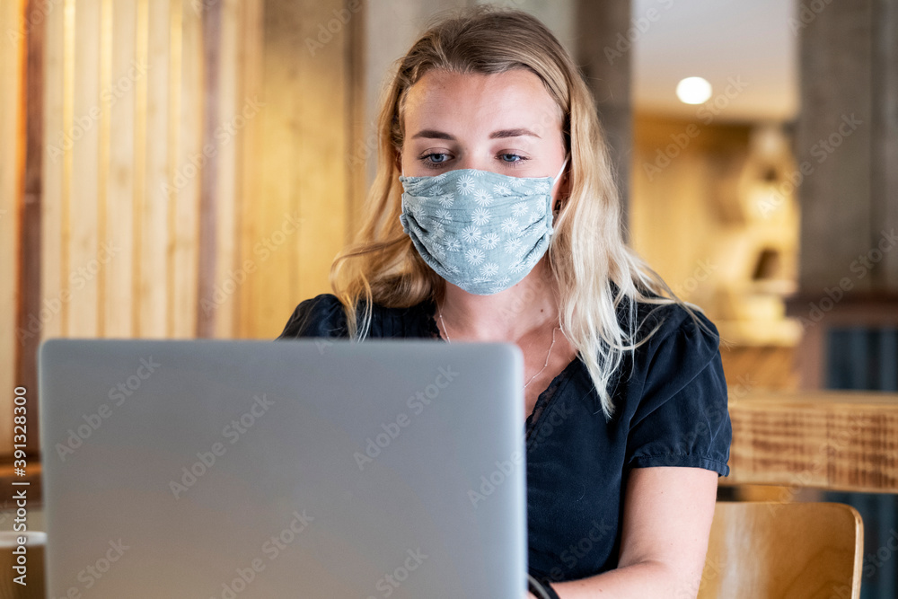Young blond woman wearing blue face mask, sitting at table, using laptop computer. Young blond woman wearing blue face mask, sitting at table, using laptop computer.