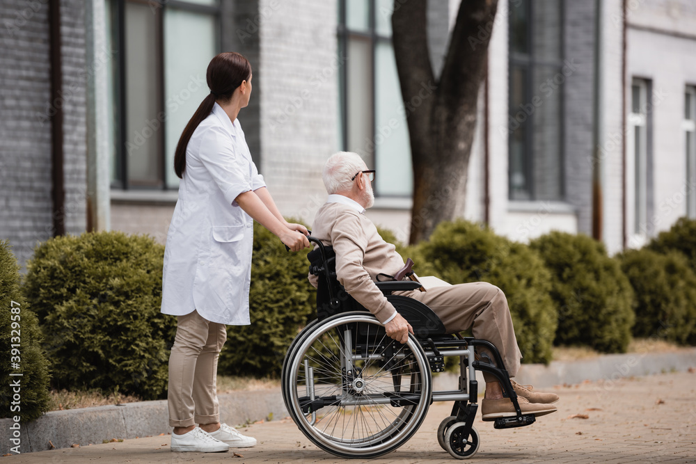 social worker walking with aged disabled man on wheelchair outdoors