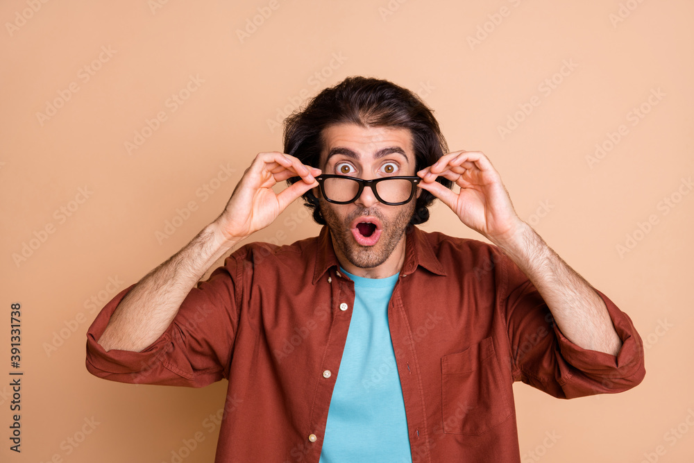 Photo of amazed young guy arms hold eyewear open mouth wear glasses brown t-shirt isolated beige color background Photo of amazed young guy arms hold eyewear open mouth wear glasses brown t-shirt isolated beige color background