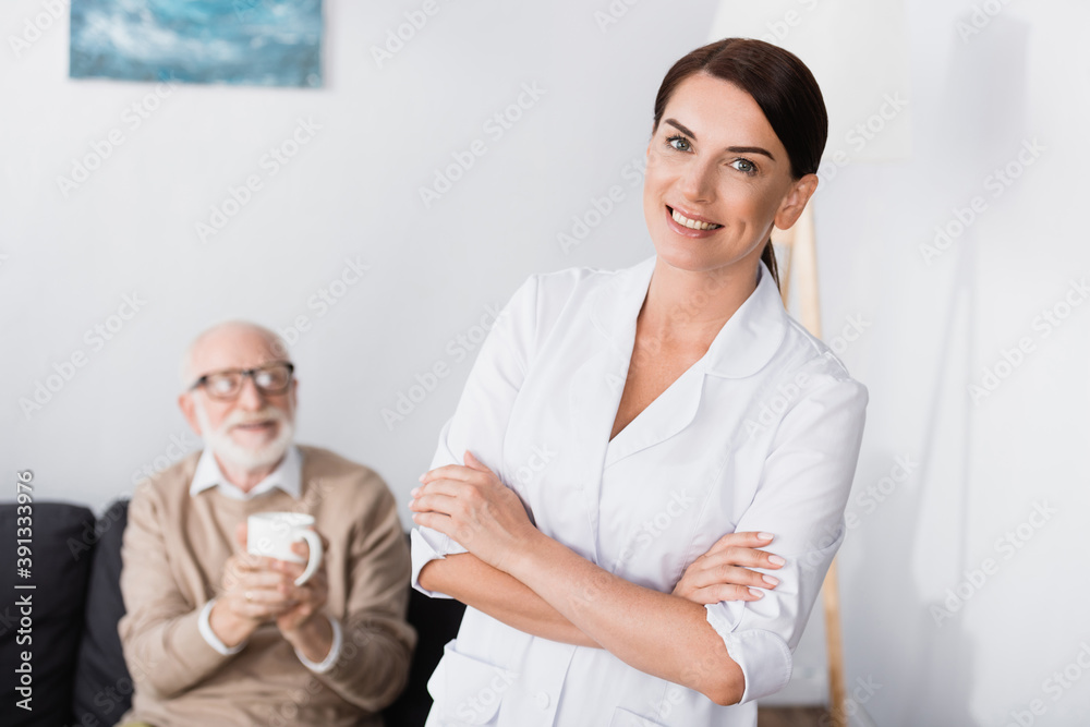 cheerful social worker smiling at camera while standing with crossed arms near aged man holding cup of tea cheerful social worker smiling at camera while standing with crossed arms near aged man holding cup of tea