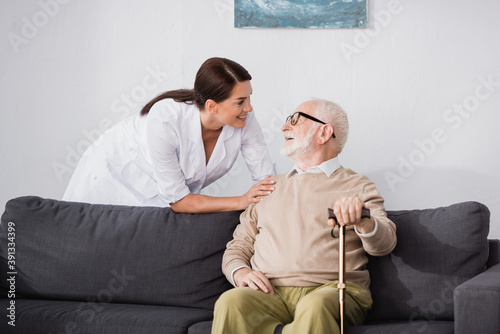 Принти на полотні smiling geriatric nurse talking to elderly man sitting on sofa at home