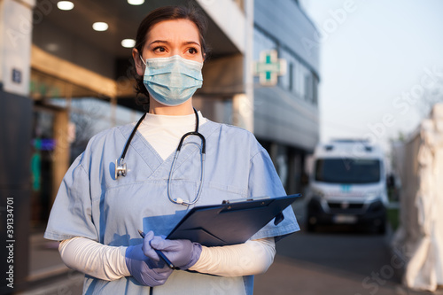 Young female EMS key worker doctor in front of healthcare ICU facility,wearing protective PPE face mask equipment,holding medical lab patient health check form,UK&US COVID-19 pandemic outbreak crisis  photo