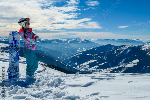 A woman posing on powder snow with her snowboard on top of Katschberg in Austria. Panoramic view on the surrounding mountains. Winter wonderland. Sunny winter day. She is full of energy and happy