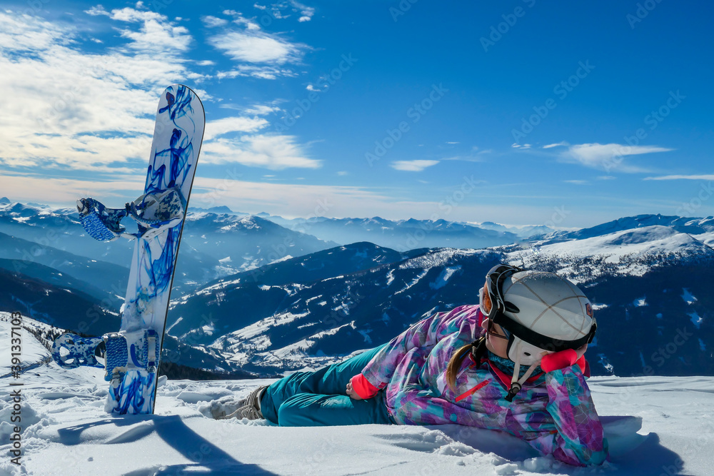 A woman lying on powder snow with her snowboard on top of Katschberg in Austria. Panoramic view on the surrounding mountains. Winter wonderland. Sunny winter day. She is full of energy and happy