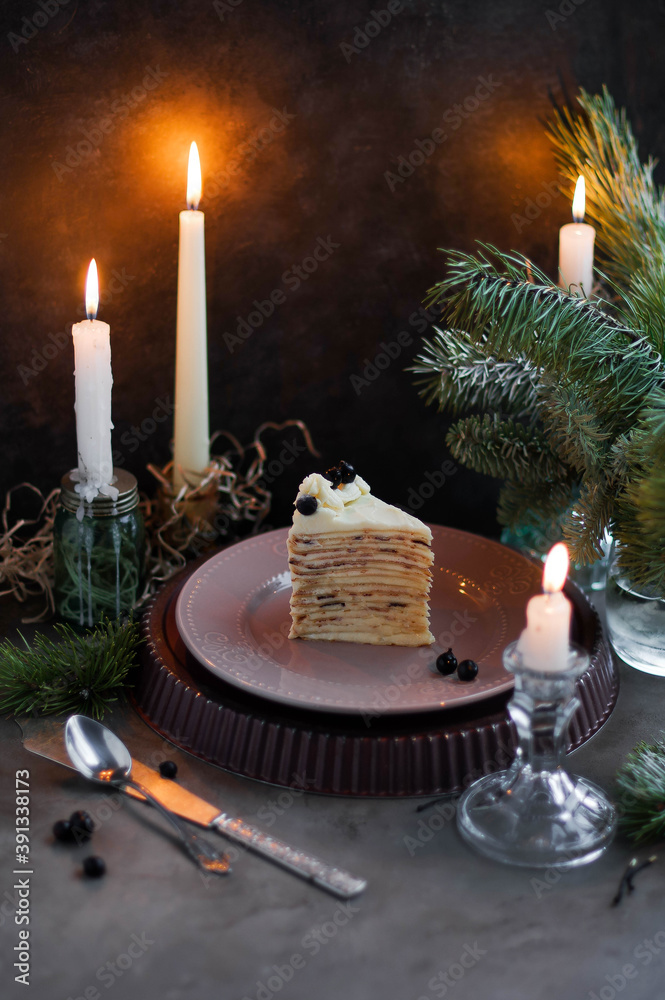 Slice of pancake cake with butter cream and black currant. Festive cake on a stand on a table with pine branches and candles. Christmas evening. Slice of pancake cake with butter cream and black currant. Festive cake on a stand on a table with pine branches and candles. Christmas evening.