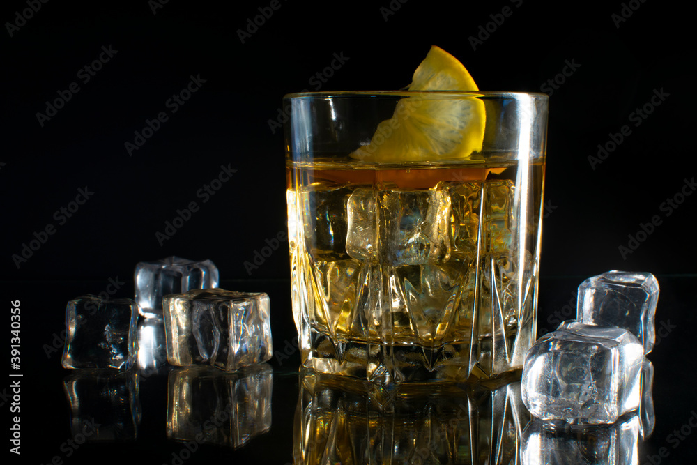 A glass of whiskey with ice with a slice of lemon on a black table with reflection. On a black background. A glass of whiskey with ice with a slice of lemon on a black table with reflection. On a black background.