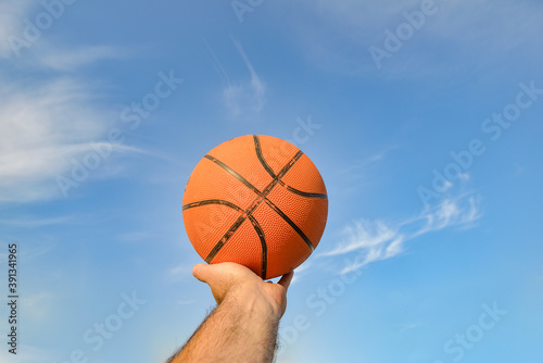 The hand of a male athlete holds a basketball ball against a blue cloudy sky. Sports activities on the playground. © andov