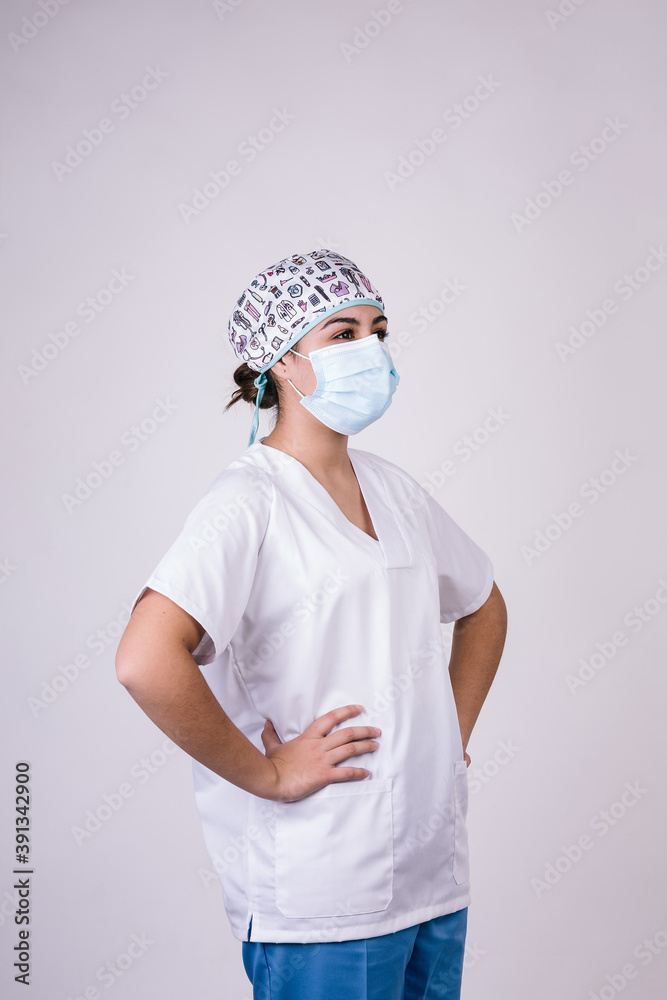 portrait of a nurse with her arms on her hips. She wears a cap and a mask for protection. The photo is made on a white background