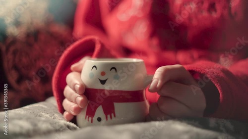 girl in Christmas pajamas drinking coffee in a cup shaped like a snowman. teenager on blue sofa warming his hands with a hot drink. hot milk in cup and hands, woman with tea.
