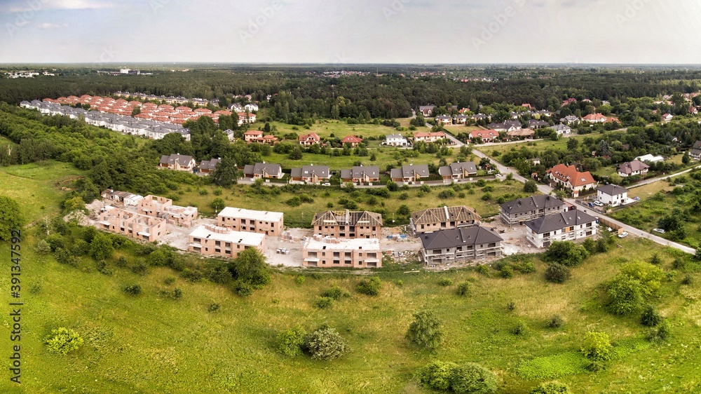 Aerial view on estate of multi-family houses under construction. House In basic state. Wooden roof structure under construction. House located on green plot in small village. Green meadows around.