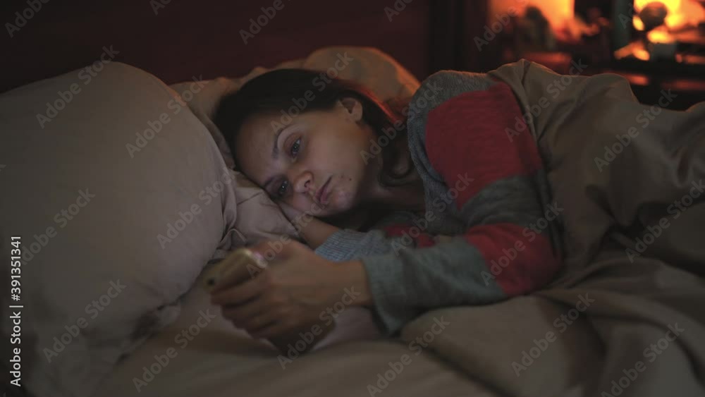 A young lonely woman lies in bed before going to bed and browses the Internet on her smartphone. A young lonely woman lies in bed before going to bed and browses the Internet on her smartphone.