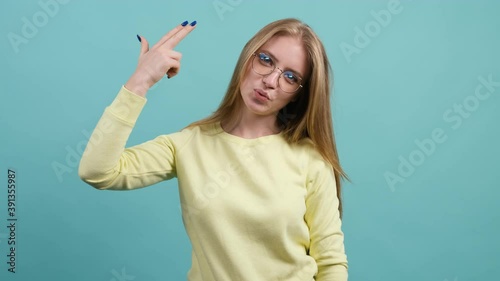 Close up of a frustrated young woman that shooting into head with finger pistol, killing herself to stop boredom, desperate emotions. isolated on turquoise background.