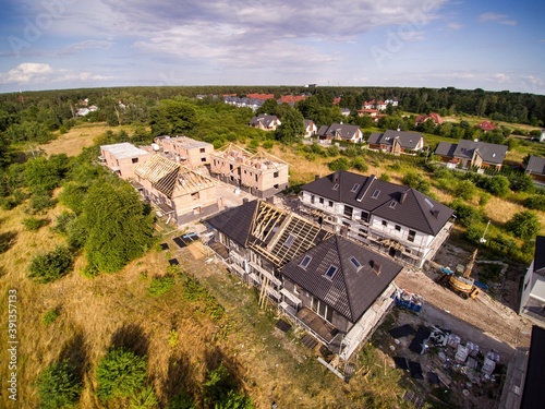 Aerial view on estate of multi-family houses under construction. House In basic state. Wooden roof structure under construction. House located on green plot in small village. Green meadows around.