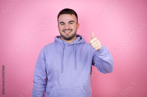 Young handsome man wearing casual sweatshirt over isolated pink background smiling and doing the ok signal with her thumb
