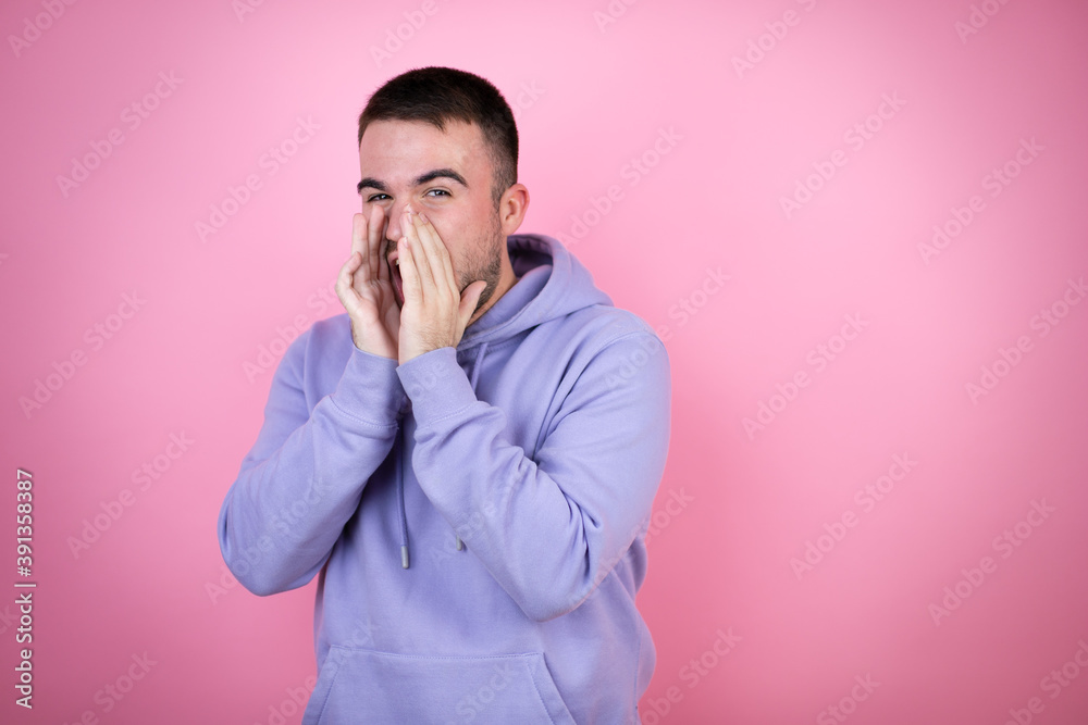 Young handsome man wearing casual sweatshirt over isolated pink background shouting and screaming loud to side with hands on mouth Young handsome man wearing casual sweatshirt over isolated pink background shouting and screaming loud to side with hands on mouth