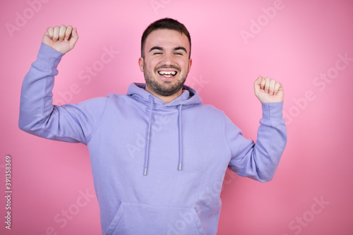 Young handsome man wearing casual sweatshirt over isolated pink background very happy and excited making winner gesture with raised arms, smiling and screaming for success. © Irene