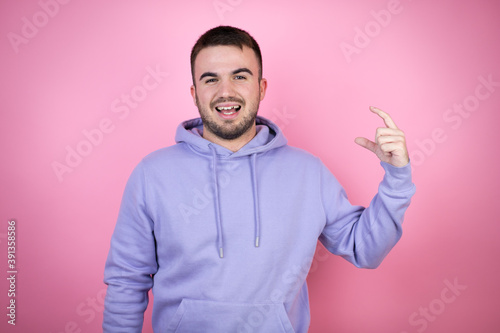 Young handsome man wearing casual sweatshirt over isolated pink background smiling and confident gesturing with hand doing small size sign with fingers . Measure concept. © Irene