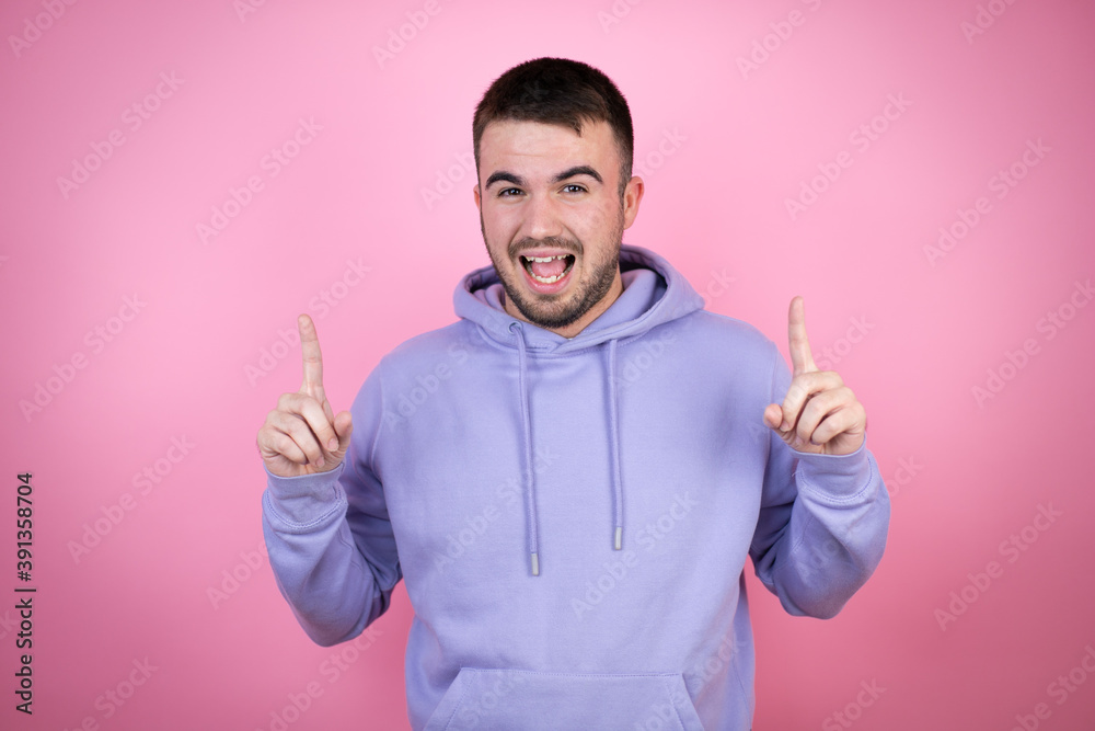 Young handsome man wearing casual sweatshirt over isolated pink background amazed and surprised looking at the camera and pointing up with fingers and raised arms