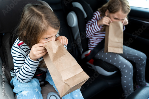 Little girls, sisters are driving in car. Children are sick, vomit into paper bag. Traveling on road in safe baby seats with child belts. Fun family trip, activity with parents photo