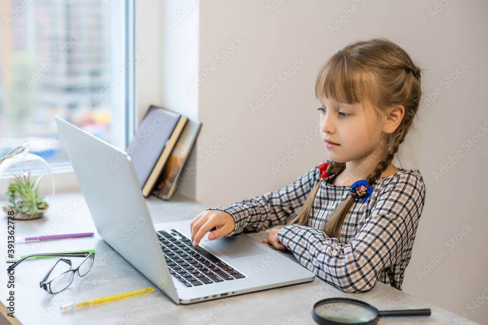 Little blonde girl doing homework at home at the table. The child is home-schooled. A girl with light hair performs a task online using a laptop and tablet computer. Little blonde girl doing homework at home at the table. The child is home-schooled. A girl with light hair performs a task online using a laptop and tablet computer.