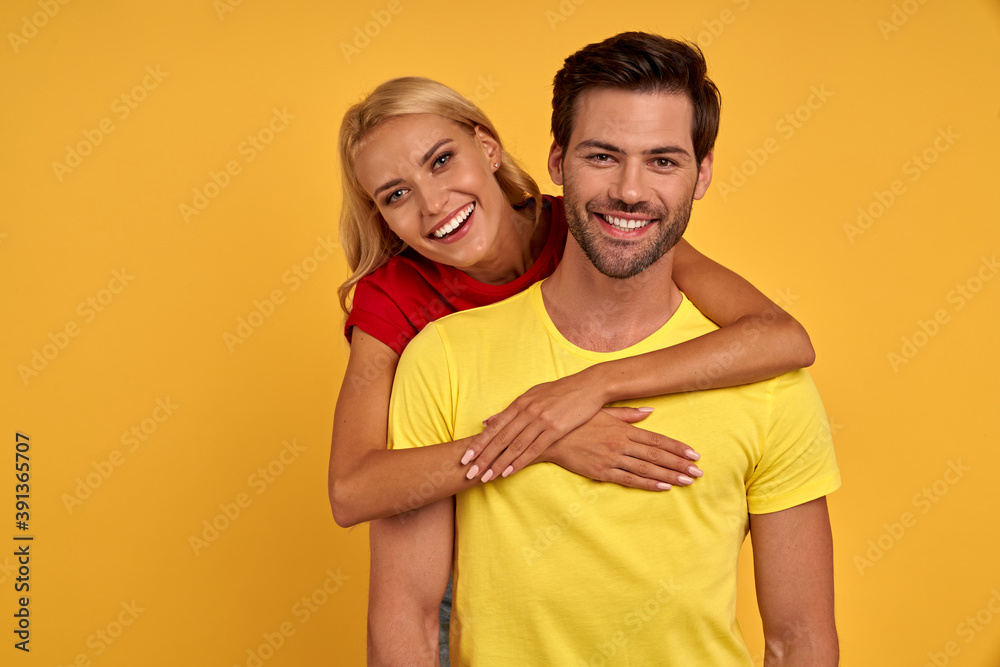 Smiling young couple friends in colored t-shirts posing isolated on yellow wall background studio portrait. People emotions lifestyle concept. Mock up copy space. Hugging Smiling young couple friends in colored t-shirts posing isolated on yellow wall background studio portrait. People emotions lifestyle concept. Mock up copy space. Hugging