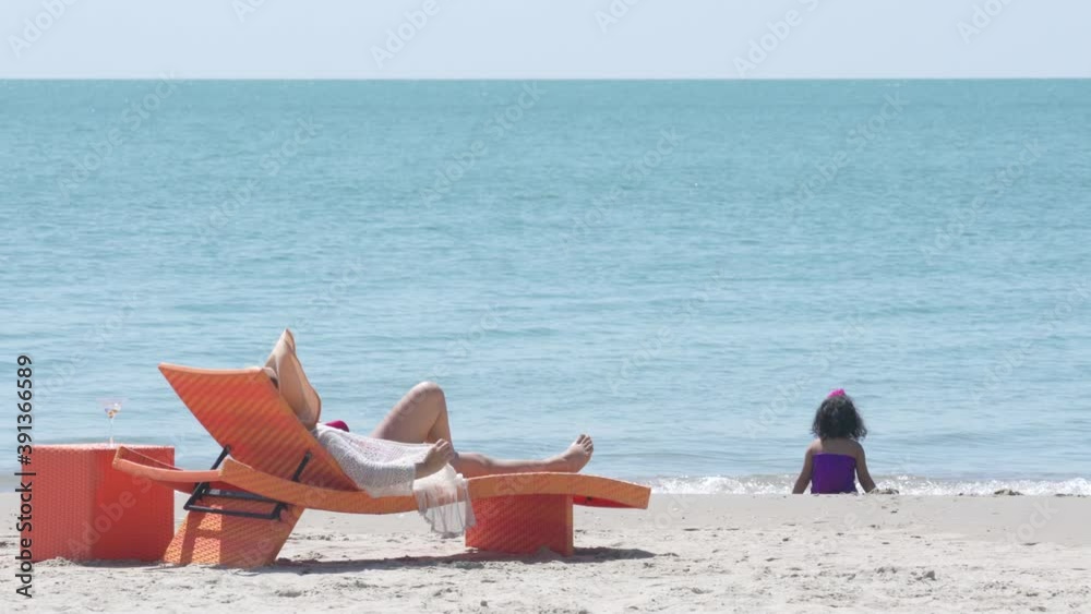 Happy Asian family holiday vacation. Asian mother resting on beach chair looking little daughter playing on the beach in summer day. Cute child girl relax and having fun together with mom on the beach Happy Asian family holiday vacation. Asian mother resting on beach chair looking little daughter playing on the beach in summer day. Cute child girl relax and having fun together with mom on the beach